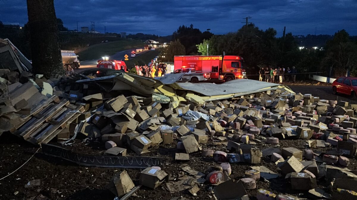 Carreta tombada com caixas de mercadorias espalhadas na BR-282, em Ponte Serrada, com viaturas dos bombeiros ao fundo